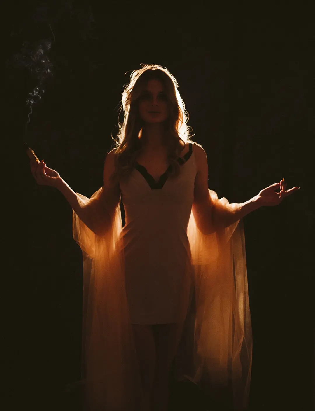 Backlit woman with sheer sleeves and outstretched arms amid rising smoke, amber light on black