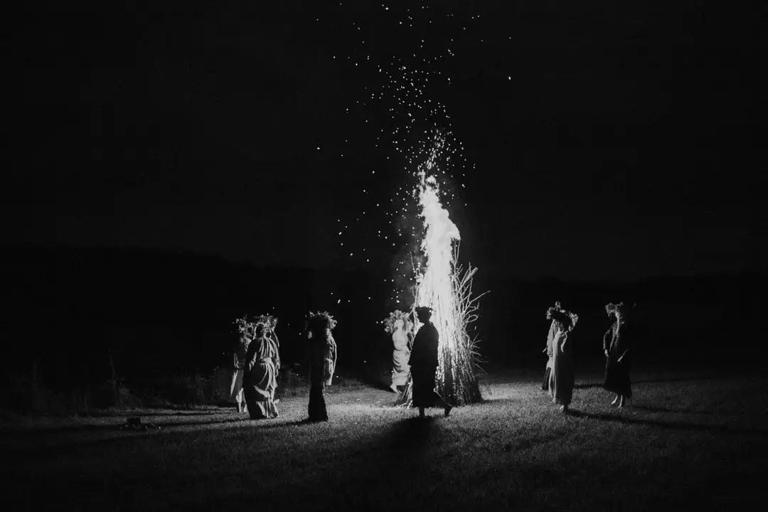 Wreath-crowned figures in long robes circling a bonfire at night, black and white