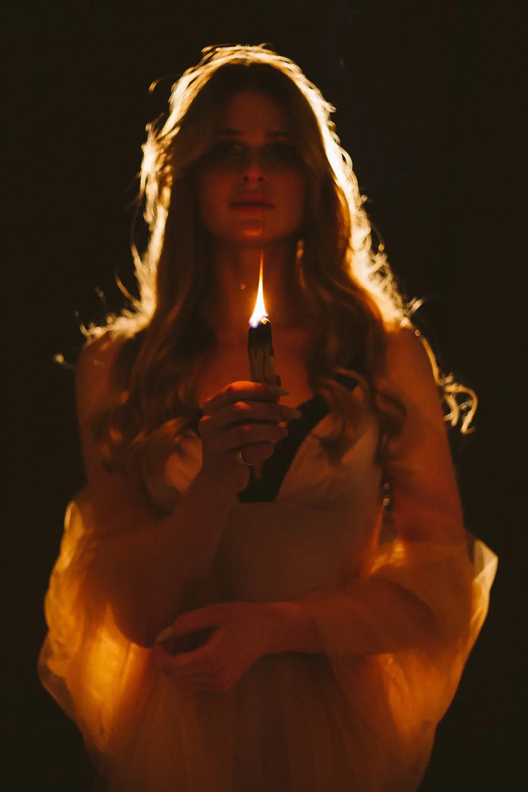Woman holding a burning stick close to her face, rim-lit hair glowing against darkness