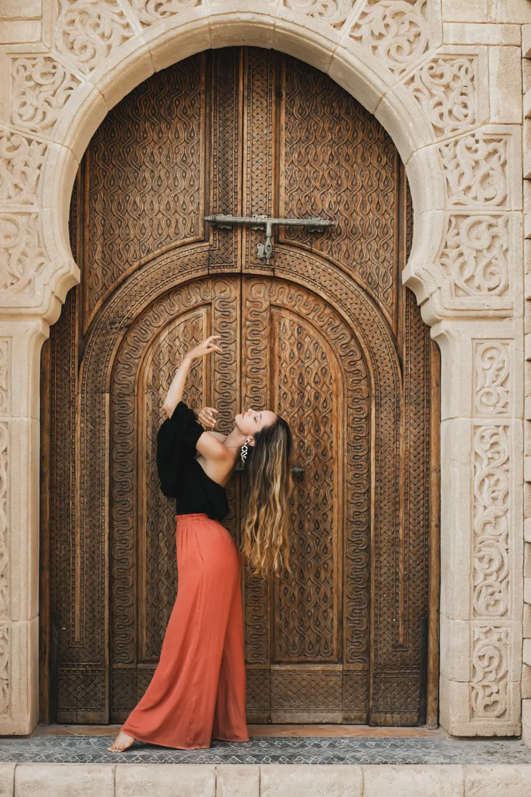 Barefoot dancer in terracotta trousers arching back before a carved wooden door and stone arch