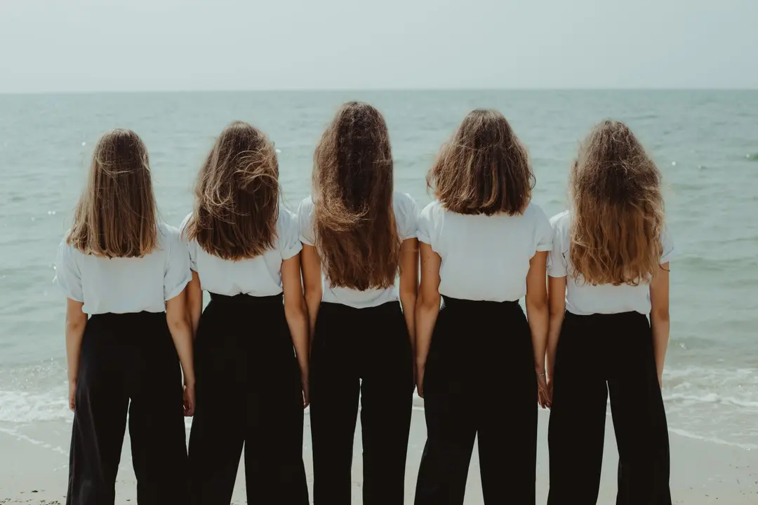 Five girls in white T-shirts and black trousers seen from behind, facing a calm sea