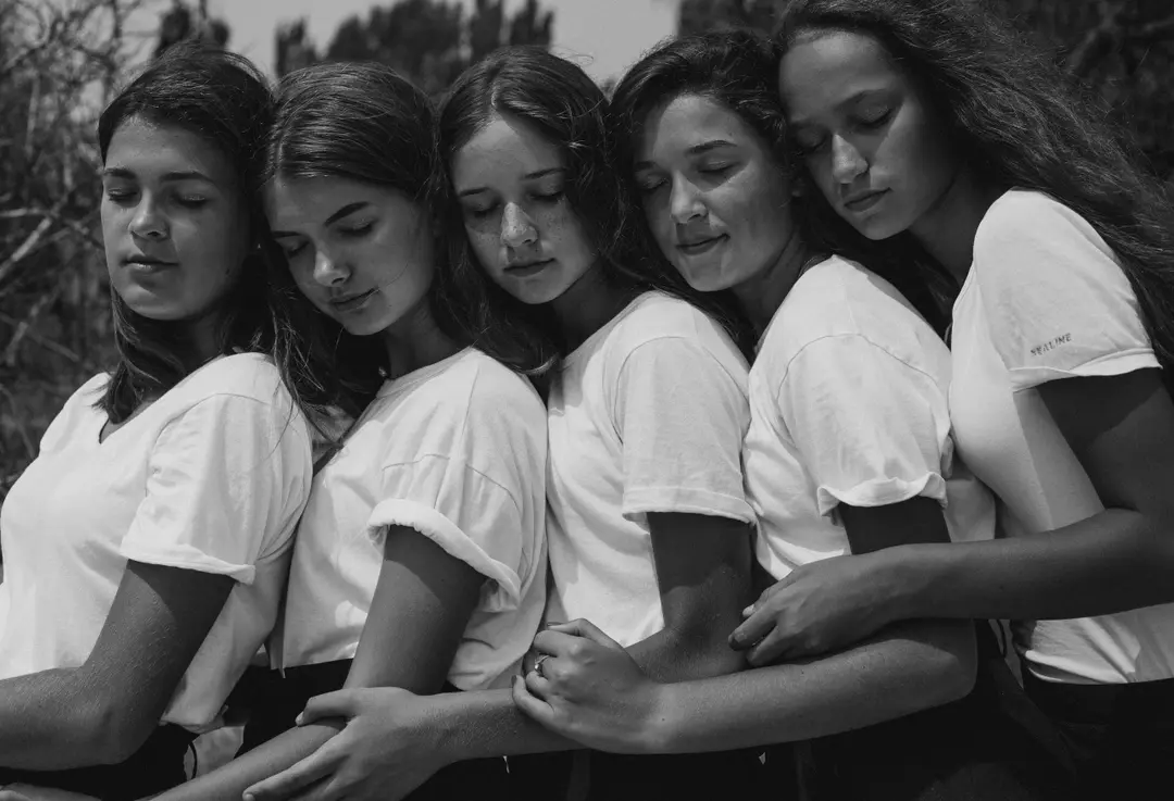 Five girls in white T-shirts lean their heads together, eyes closed, black and white