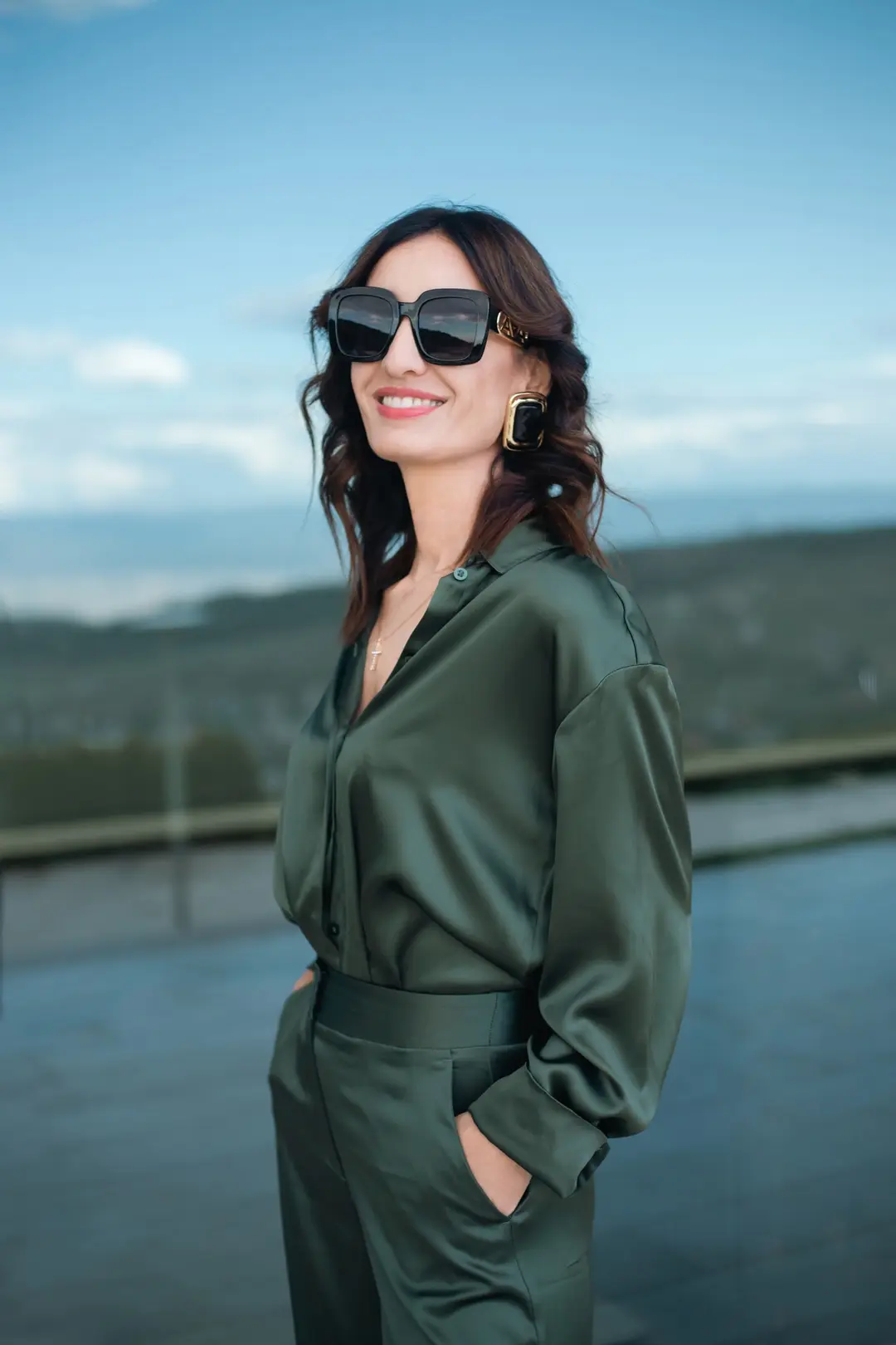 Smiling woman in a green satin shirt and square sunglasses on a terrace above green hills