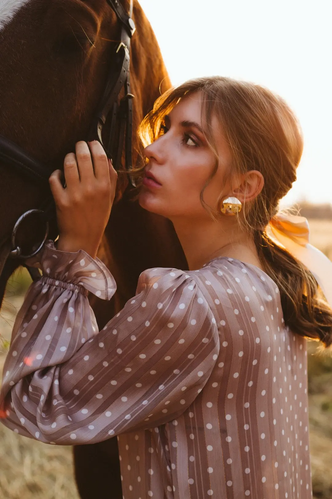 Woman in a polka-dot blouse kissing a chestnut horse's muzzle in golden backlight