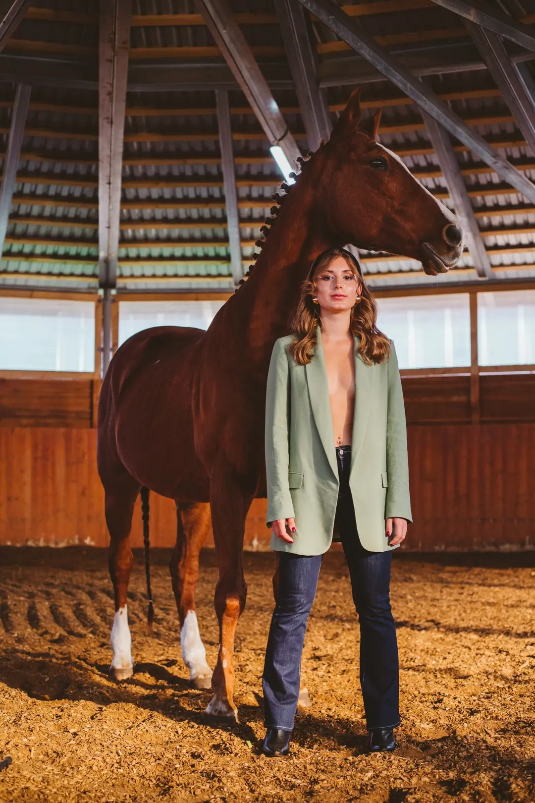 Woman in a sage blazer and jeans beside a chestnut horse in a wooden indoor arena