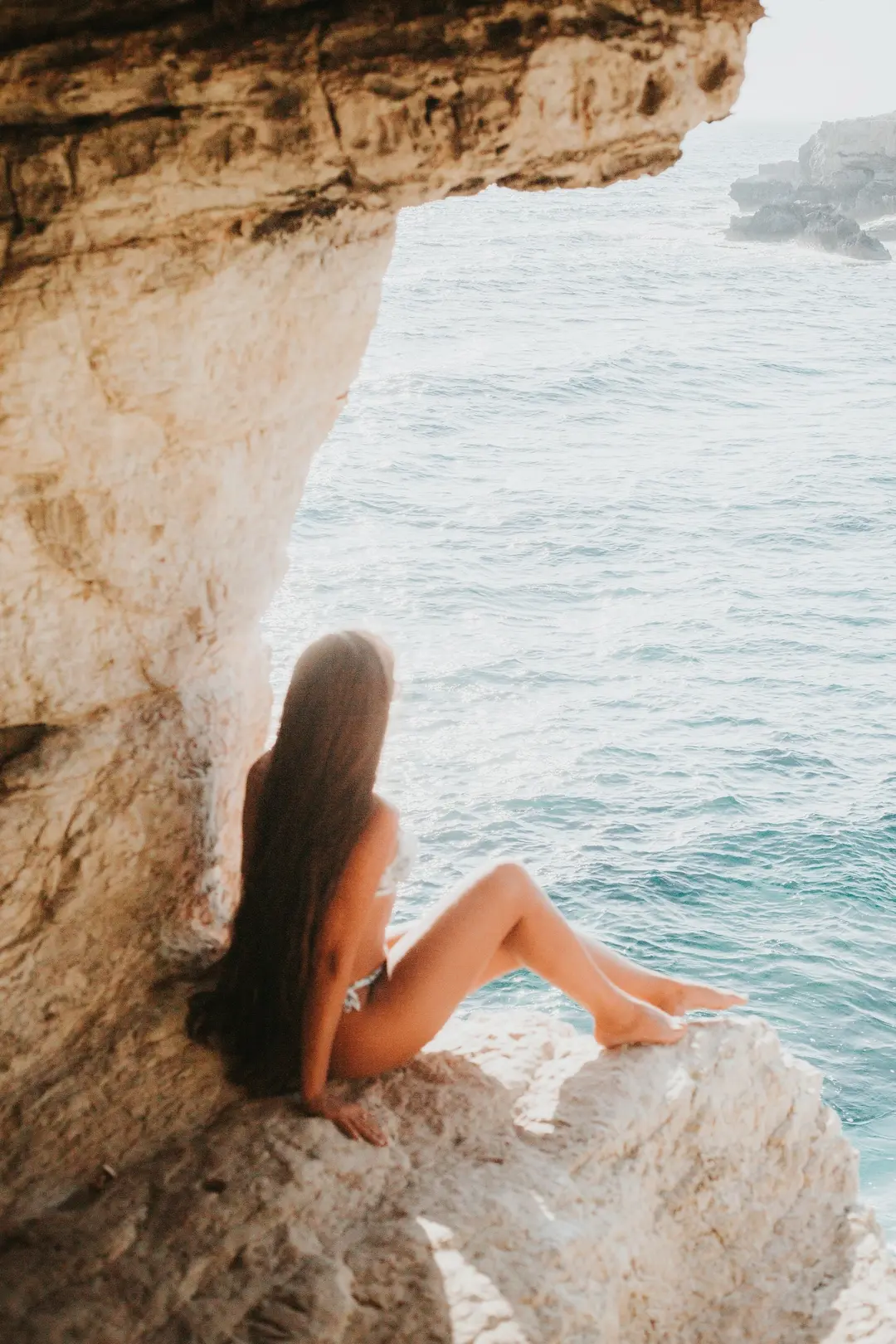 Long-haired woman seen from behind, seated in a sea-cave arch above turquoise water