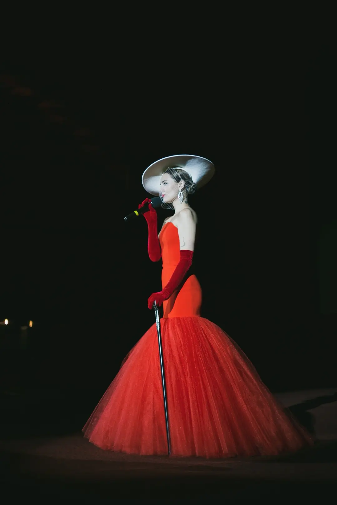 Singer in a red tulle gown and wide-brimmed hat, seen in profile on a dark stage