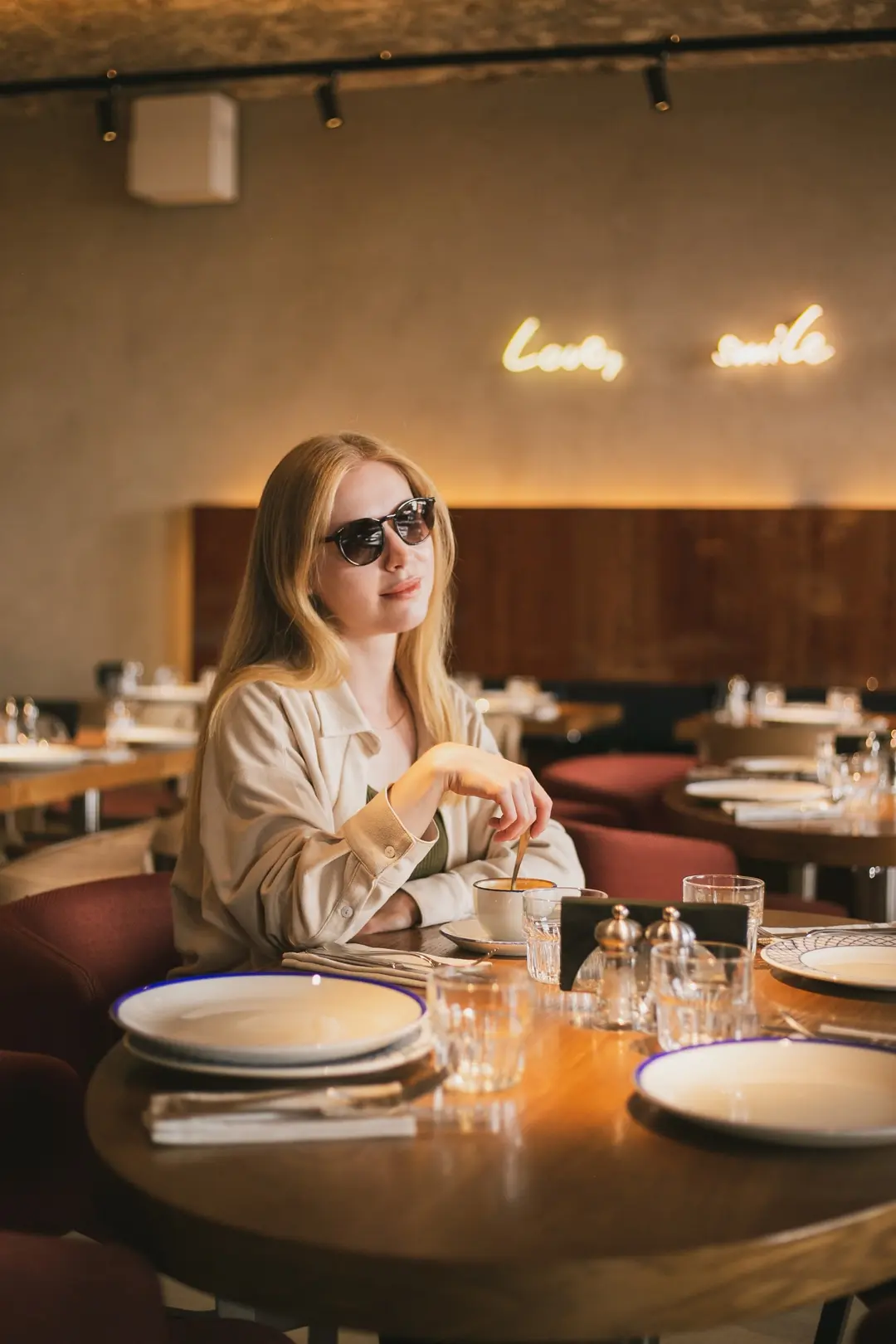 Woman in sunglasses with coffee at a restaurant table beneath glowing neon script signs