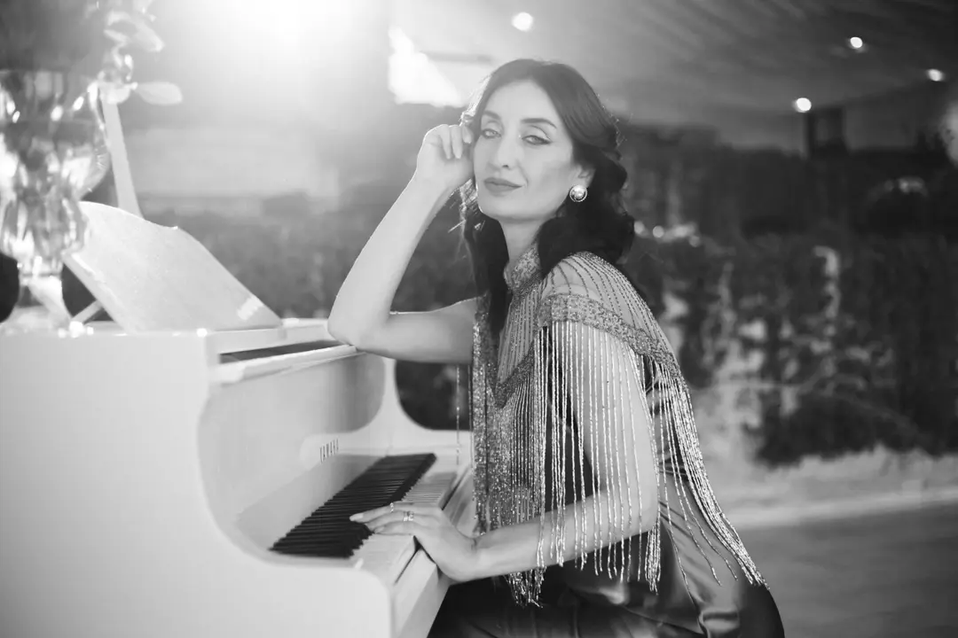 Black and white portrait of a woman in a fringed cape leaning on a white piano